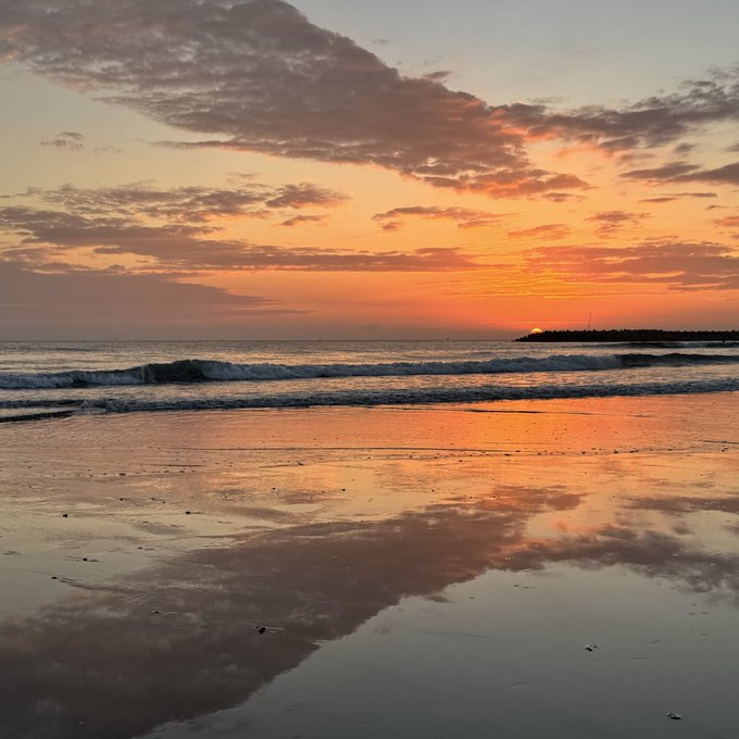 A serene sunset over the ocean, with orange and pink hues reflected on the wet sand of a beach.