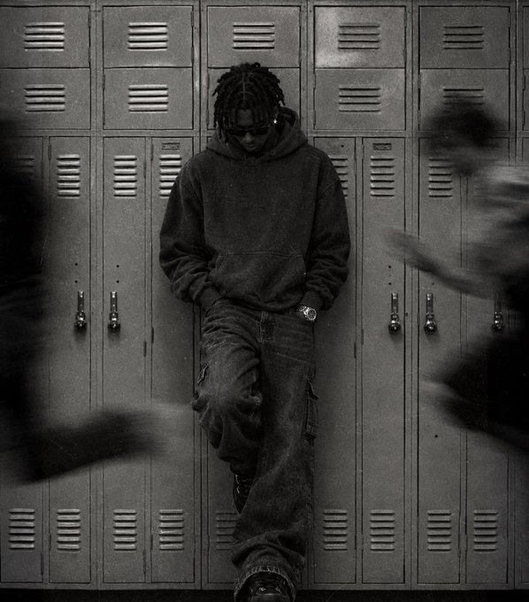 A black-and-white photo of a young man with dreadlocks, sunglasses, and a hoodie leaning against lockers while blurred figures rush past in the foreground.