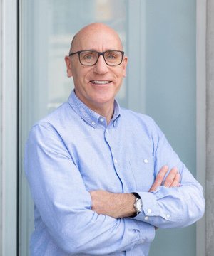 A professional headshot of a smiling bald man wearing glasses and a light blue button-down shirt with his arms crossed.