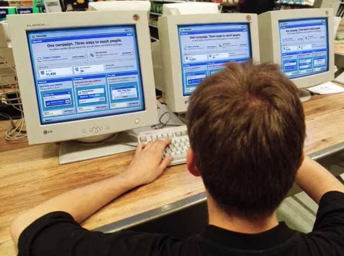 A young boy with short brown hair sits at a wooden desk using a white LG Flatron monitor displaying the Facebook homepage with 'One campaign. Three ways to reach people.'
