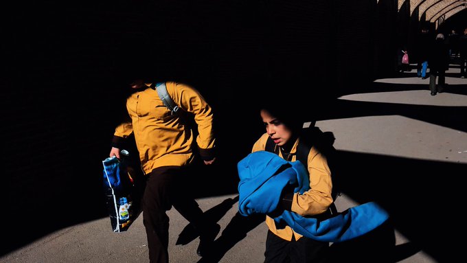 Two people wearing yellow jackets are walking in a dimly lit outdoor area, with one carrying a bright blue backpack and the other pulling a suitcase.
