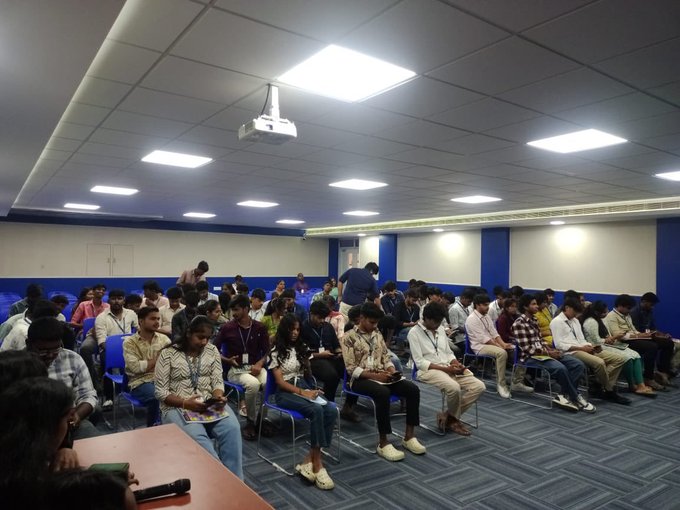 A crowded conference room or classroom with seated attendees facing forward, featuring a ceiling projector, blue and white walls, and fluorescent lighting.