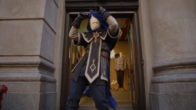 A person in a blue and white bird-like mascot costume standing in a doorway, wearing a dark robe with mystical symbols.