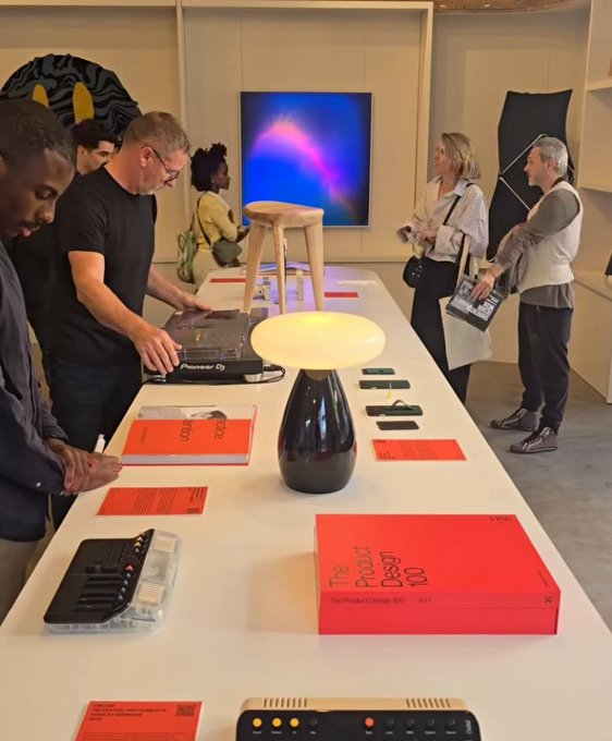 An interior photo of a design exhibition where visitors browse products on a long white table, including a wooden stool, a mushroom-shaped lamp, and red books labeled "homo design 03."