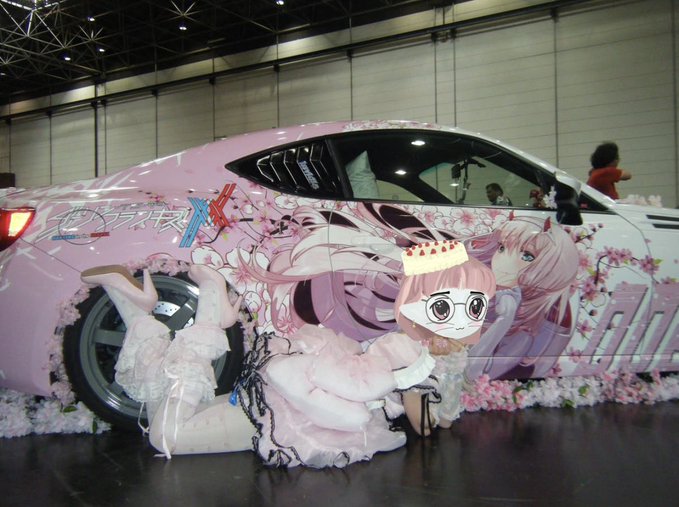 A pink anime-themed decorated car with 'ChouCho X' text parked at what appears to be a convention, accompanied by a cosplayer in a pink outfit with chains.
