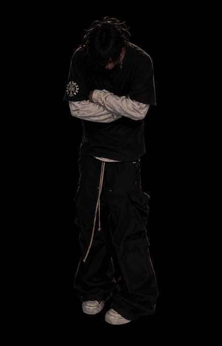 A person with long dark hair stands against a black background with their head bowed and arms crossed, wearing a black t-shirt with a small white logo, layered over a distressed long-sleeve shirt, and oversized black cargo pants with light sneakers. The image reinforces the post's mood of quiet anticipation, mirroring the introspective tone of waiting to see what gets revealed tomorrow from @EthraShip.