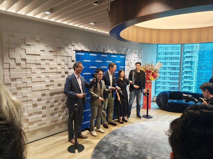 A corporate ribbon-cutting ceremony at a Coinbase-branded event, showing five people standing together cutting a red ribbon in a modern office space with a flower arrangement and large windows.