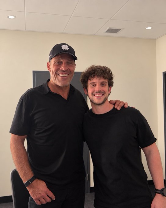 A photograph of two smiling men posing together indoors; the older man wears a black polo and a Clemson cap, while the younger man has curly hair and wears a black t-shirt.