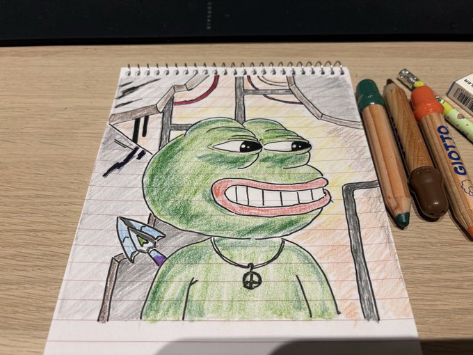 A hand-drawn colored pencil sketch of the Pepe the Frog meme with a smug grin, drawn in a spiral notebook next to a collection of colored pens and markers on a wooden desk.
