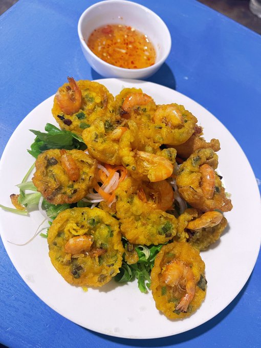 A white plate of golden Vietnamese-style shrimp and vegetable fritters served over greens with shredded carrots, accompanied by a bowl of fish sauce-based nuoc cham dipping sauce, all on a blue table.