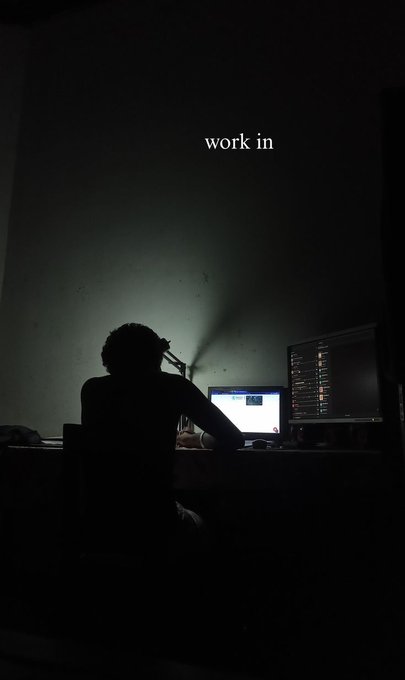 A silhouetted figure works at a desk with dual monitors in a dark room, illuminated only by the screen glow, with the text "work in" overlaid in white.