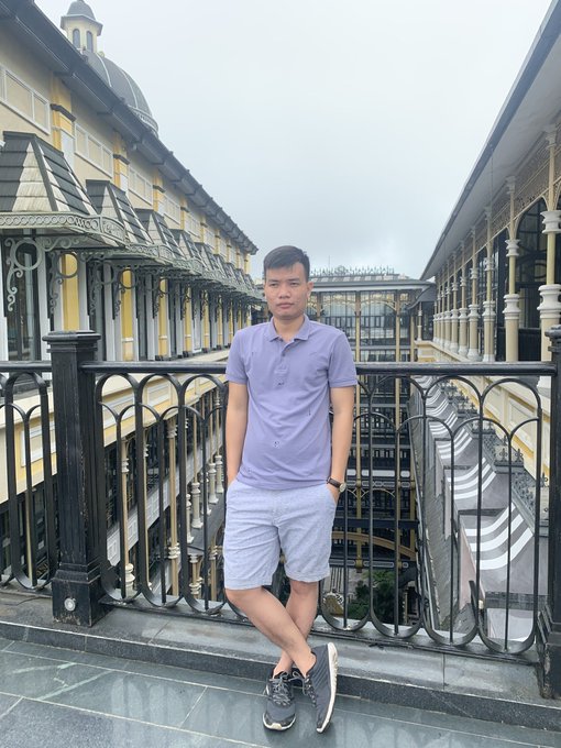 A man in a lavender polo shirt and gray shorts stands on a balcony of an ornate building overlooking an indoor shopping arcade.