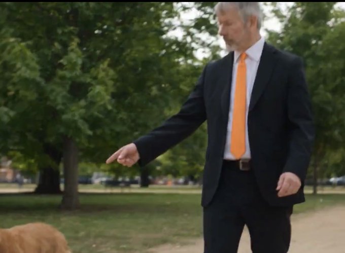 A man in a dark suit with a bright orange tie points his finger downward toward a dog partially visible in the lower left corner of the park setting, humorously illustrating the post's joke about Michael Saylor acknowledging Bitcoin (DOGE) by literally pointing at a dog.