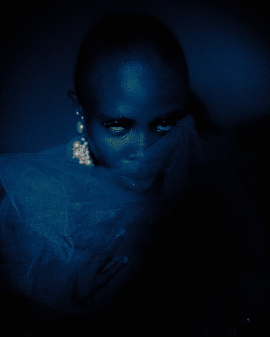 A dark, atmospheric portrait of a woman illuminated by blue light, wearing pearl earrings and a sheer garment, with a moody and mysterious expression.