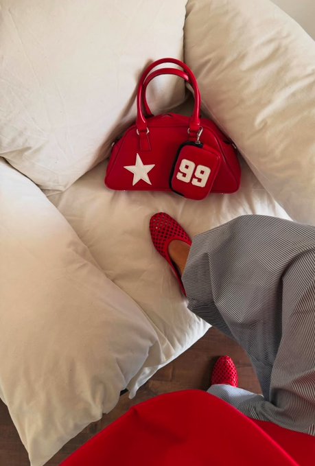 The image shows a pair of red shoes with white stars on them placed next to a purse that has the number 99 on it. The purse appears to be made of leather and is positioned near a couch cushion. The background features a gray wall, which contrasts with the vibrant colors of the shoes and purse.