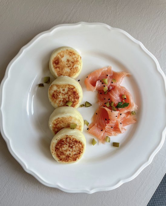Four golden-brown potato syrniki are arranged beside delicate slices of smoked salmon, garnished with chopped pickles and herbs on a scalloped white plate, paired with a "potato syrniki #cryptosoupgroup" caption that highlights a creative fusion of traditional Eastern European pancakes with modern plating.