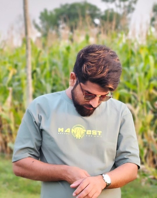 A bearded man wearing sunglasses and a sage green t-shirt with 'M4 BST' text, standing in a cornfield and looking at his watch.