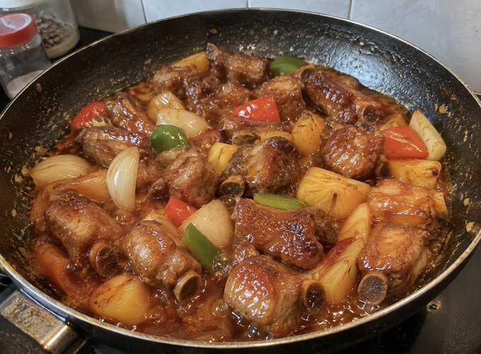 A pan of braised pork ribs simmering in a dark savory sauce with onions, bell peppers, and pineapple chunks on a stovetop.