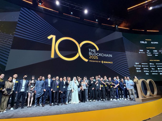 A large group of attendees poses on stage at 'THE BLOCKCHAIN 2025' event presented by Binance, featuring a large '100' logo and sponsor displays for CELO and Lumia.