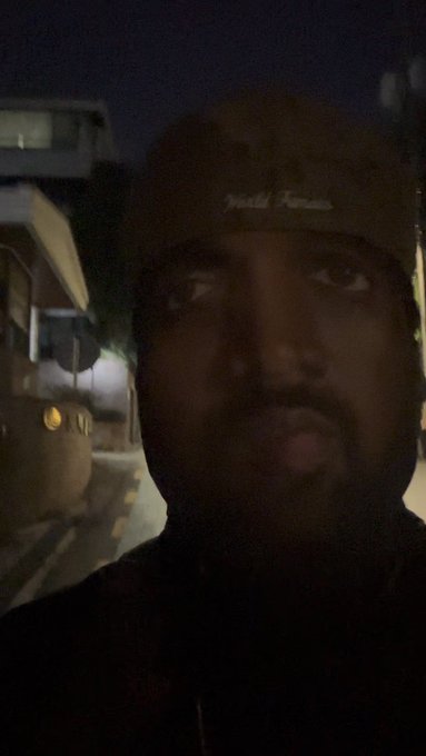 A dark, low-quality nighttime selfie of a bearded man wearing a beanie, taken outdoors on a street with a parked vehicle and building visible in the dim background.
