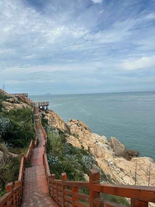 A wooden boardwalk winds along a rocky coastal cliff overlooking the ocean, with a distant island visible on the horizon under a partly cloudy sky.