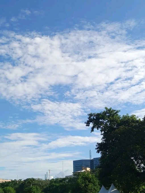 Bright blue sky with scattered white clouds over a cityscape featuring trees, a construction crane, and white tent structures.