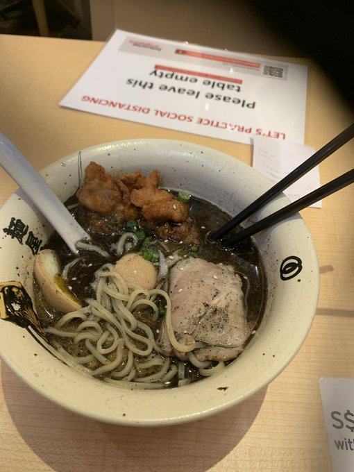 A bowl of ramen with noodles, meat, and a halved boiled egg, placed on a table next to a notice that reads "LET'S PRACTICE SOCIAL DISTANCING Please leave this table empty" upside down.