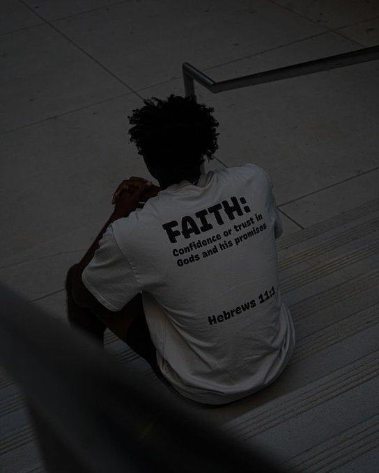 Person with curly dark hair sitting on concrete steps, viewed from behind, wearing a white t-shirt with the text 'FAITH: Confidence or trust in Gods and his promises - Hebrews 11:1'.
