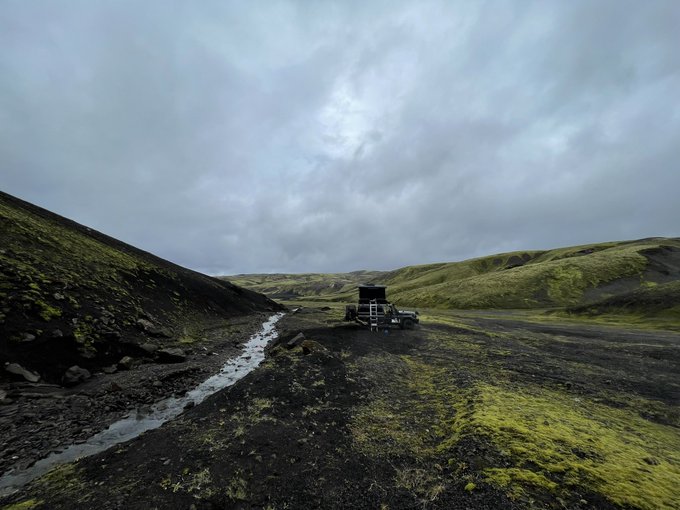 A moody, overcast landscape shows a rugged terrain with dark volcanic soil, patches of green moss, a small stream, and a vehicle or trailer parked in the middle distance.