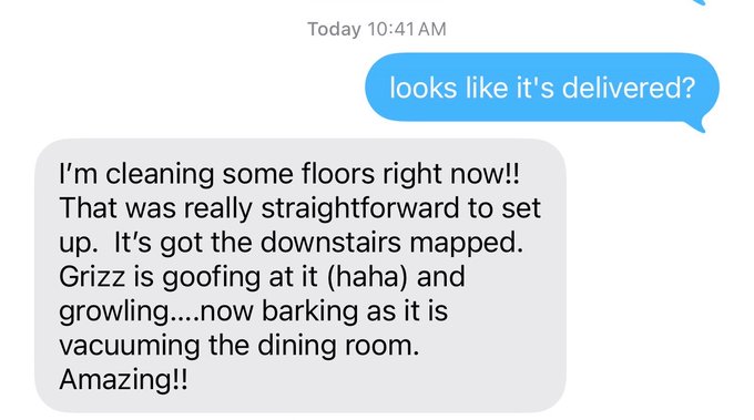 A text message conversation where one person asks "looks like it's delivered?" and the other responds excitedly about a robot vacuum mapping the downstairs and their dog "Grizz" barking at it while it cleans.