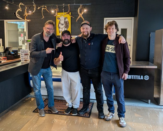 Four men posing together indoors in what appears to be a restaurant or bar, with antler decorations and a yellow banner with a goat logo visible behind them.