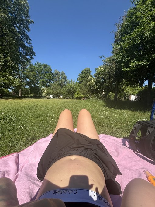 The image shows a person lying down on a blanket in a park or field with trees in the background. The person appears to be wearing a swimsuit, indicating that they are likely enjoying a day at the beach or pool. The photo has been captioned "Happy @AbstractChain XP day, guess my XP in the comments." This suggests that the image was taken on a special occasion related to the post's content.