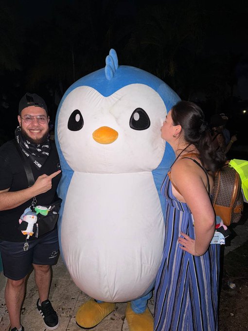 Two people posing with a large inflatable penguin mascot at night, with one person pointing at it and the other kissing its cheek.