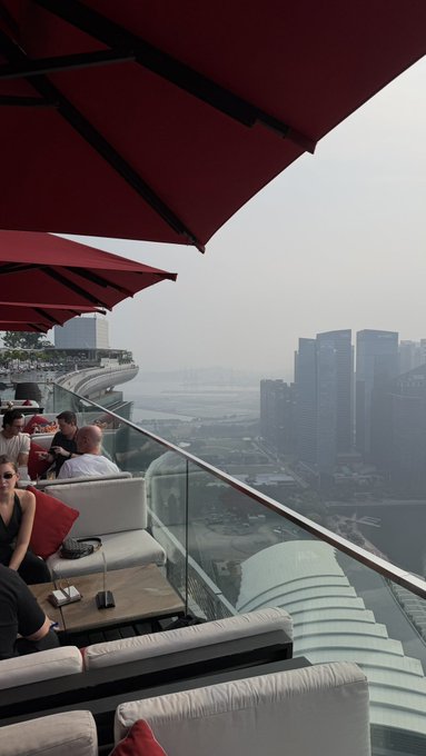 a view from a rooftop lounge with red umbrellas overlooking a hazy city skyline and body of water.