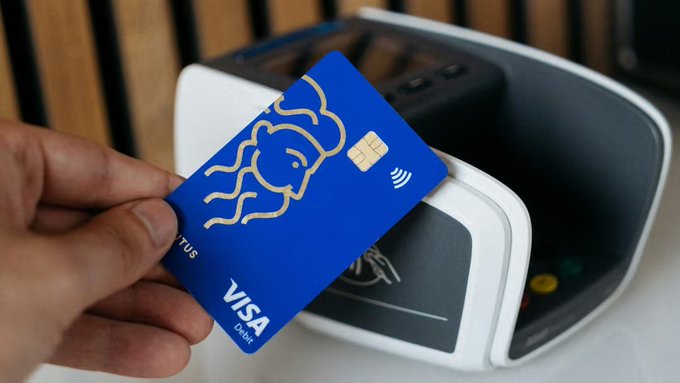 A hand holds a blue Visa Debit card featuring a stylized face design and contactless chip symbol near a payment terminal.