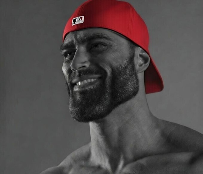 A black-and-white portrait of a smiling, bearded muscular man wearing a red MLB baseball cap backwards.