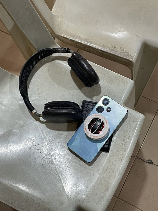 A photograph of a black pair of over-ear headphones resting on a white stone bench next to a smartphone with a light blue case featuring a phone grip holder.