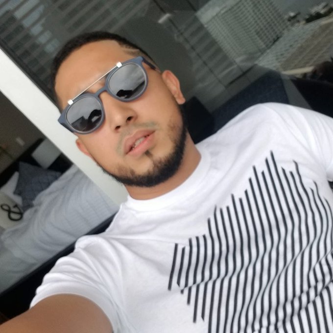 A selfie of a young man with a beard wearing dark sunglasses and a white striped t-shirt, taken in front of a window showing a city skyline.