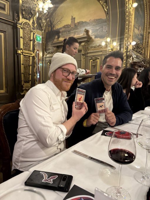 Two smiling men wearing business attire sit at a fancy restaurant holding up Pokemon cards, with a woman server in the background and a glass of red wine on the table.