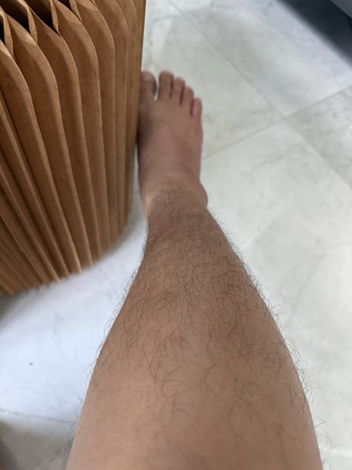 A person's leg, noticeably unshaven with visible hair growth, rests next to a wooden slatted stool on a tiled floor. The image humorously visualizes the term "soft shilling" referenced in the post about Asia markets and $TOESCOIN, playing on the wordplay between "shilling" (the currency) and "shaving" (referencing the unshaven leg).