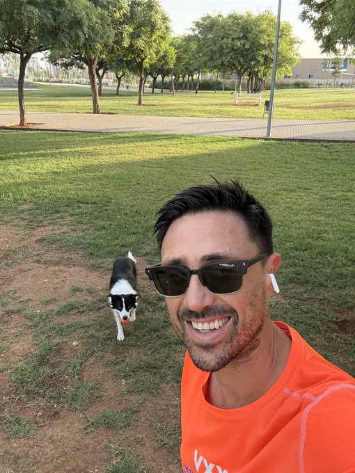 A smiling man wearing sunglasses, a bright orange shirt, and white earbuds takes a selfie in a park while a black and white dog stands on the grass behind him.
