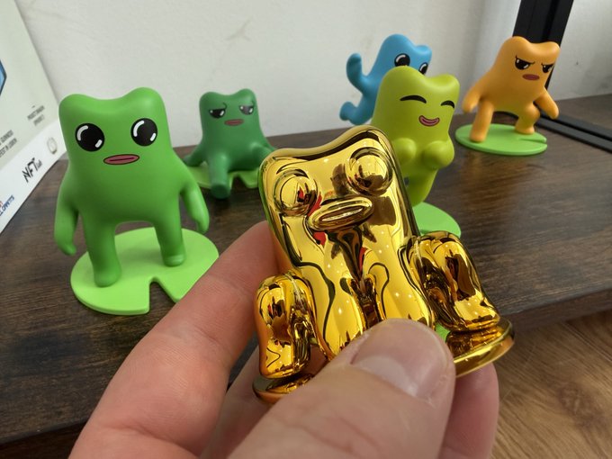 A hand holds a small shiny gold figurine in the foreground, with several colorful cartoon-style rubber figures (green, blue, and orange) displayed on a wooden surface behind it.