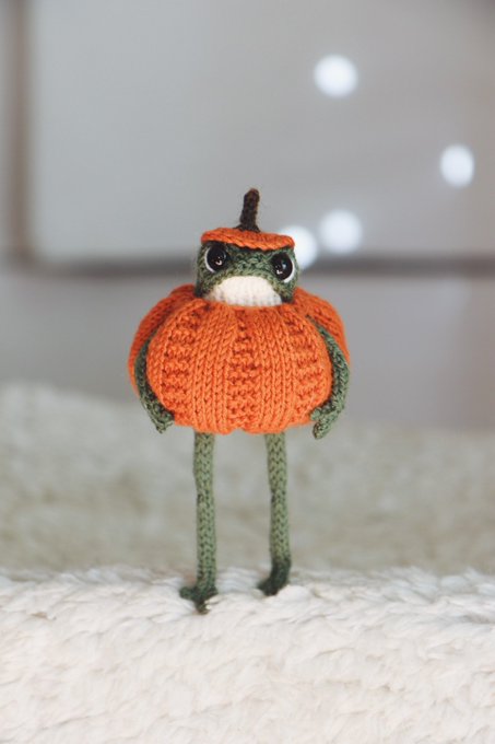 A photograph of a small, knitted amigurumi figure resembling a frog wearing a pumpkin costume, standing on a textured white blanket.
