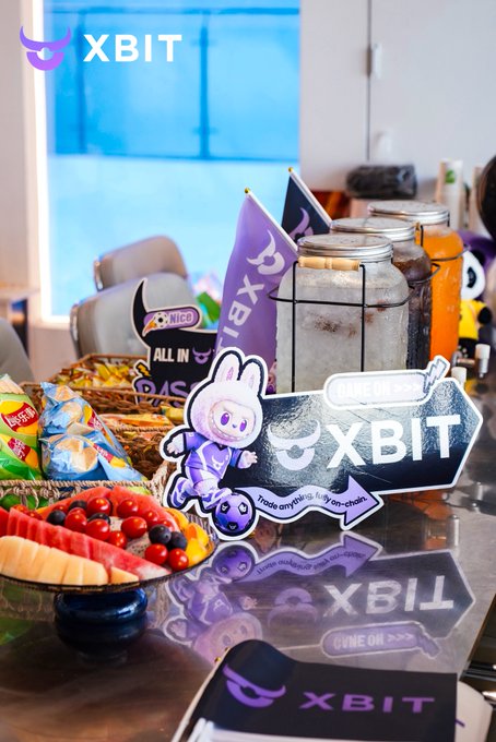 A spread of fruit platters and snacks is displayed on a table with "XBIT" branded cutouts and a small purple mascot, alongside a glass jar and orange beverages, reflecting a casual networking setting. The post thanks G+Labs and XBITDEX for hosting the enjoyable Shenzhen event, where attendees enjoyed food and connected with various crypto community members.
