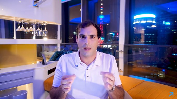 A man in a white polo shirt sitting in front of a window with a city skyline at night, gesturing with his hands while speaking.