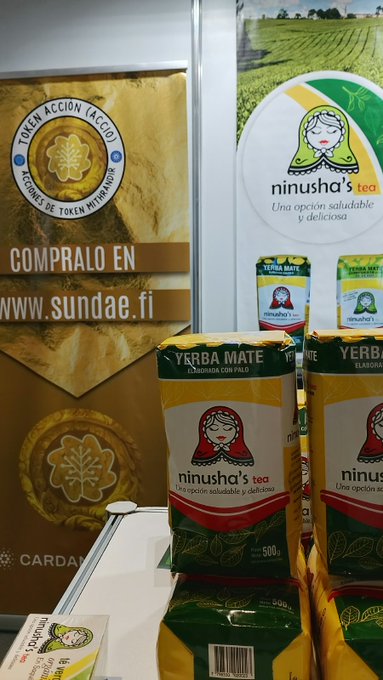 Display of 'ninusha's tea' yerba mate packages (Yerba Mate Elaborada con Palo) at a trade show booth, with a banner for the sundae.fi 'TOKEN ACCIÓN (ACCIO)' token sale.