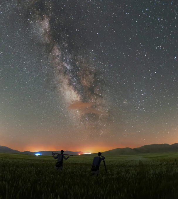 Two silhouetted figures with camera equipment stand in a grassy field under a vivid Milky Way galaxy, with warm city lights glowing on the distant horizon.