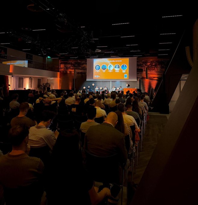 Attendees fill a dimly lit conference hall at Dutch Blockchain Week, watching a panel on a stage with a large screen displaying "MEET THE SPEAKERS" and headshots. The image captures the packed audience and stage setup, reflecting the post's celebration of Yieldbay's 2nd place win at the Solana Demo Day and the networking opportunity at the SuperteamNL-organized event.