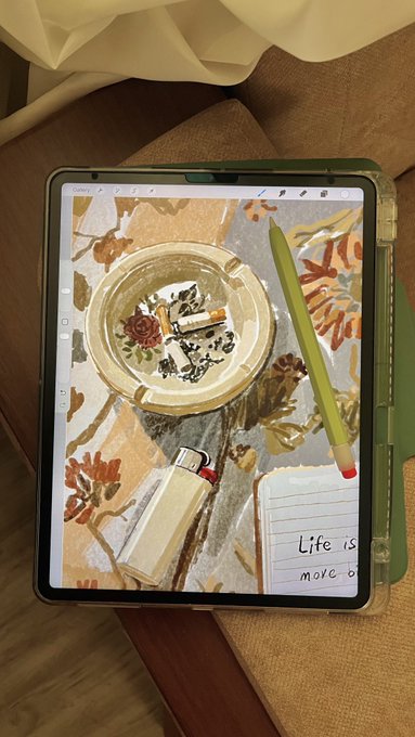 A foldable tablet rests on a brown cushion, displaying a digital painting of an ashtray with a rose design on a floral patterned surface, with a green stylus laying across the screen and a notepad reading "Life is more o..." visible.