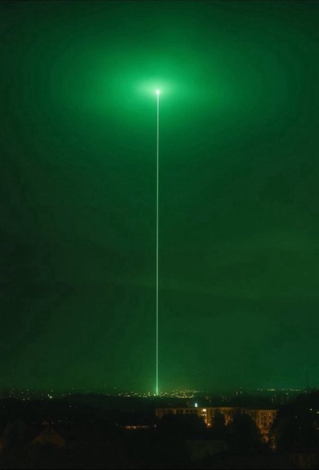 A nighttime photograph of a cityscape with a bright vertical green laser beam shooting up into the dark sky from a rooftop.